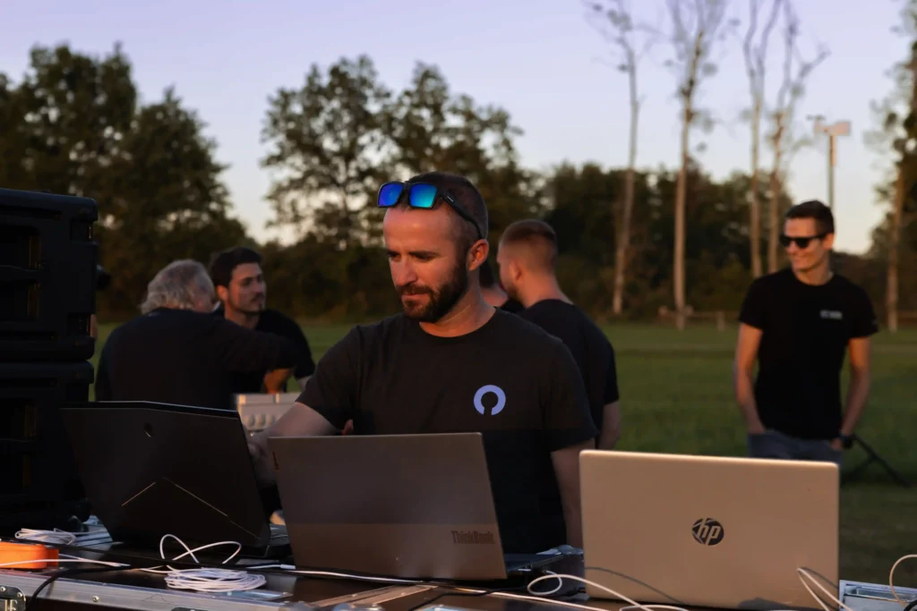 A group of technicians operating laptops and equipment outdoors at sunset, preparing or managing a drone light show in a grassy field.