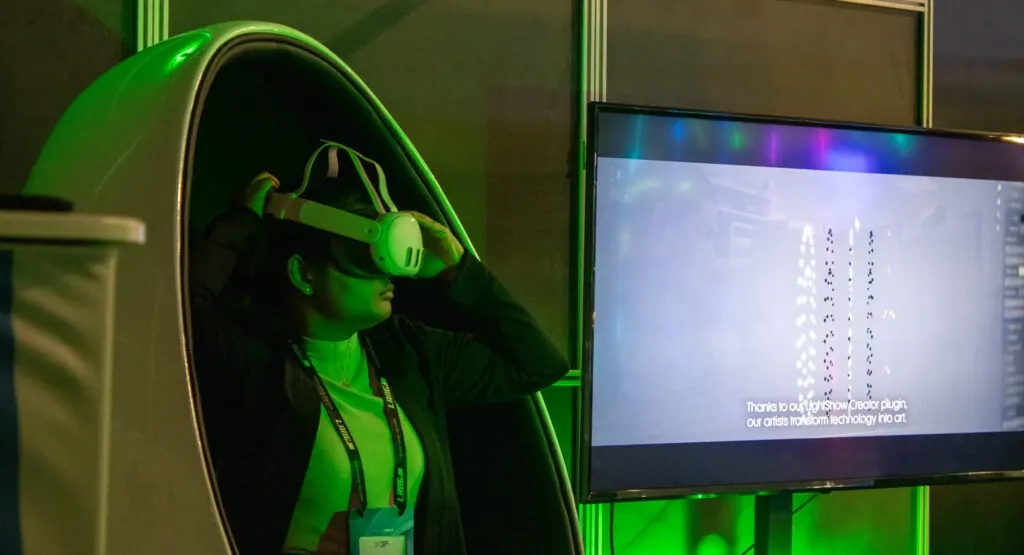 Person using a VR headset inside a booth pod, experiencing a virtual drone light show while a nearby screen displays the simulation.