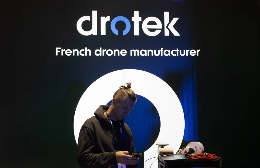 Man standing at a Drotek exhibition booth, looking at his phone in front of a large illuminated logo and the text “French drone manufacturer,” with drone equipment displayed nearby.