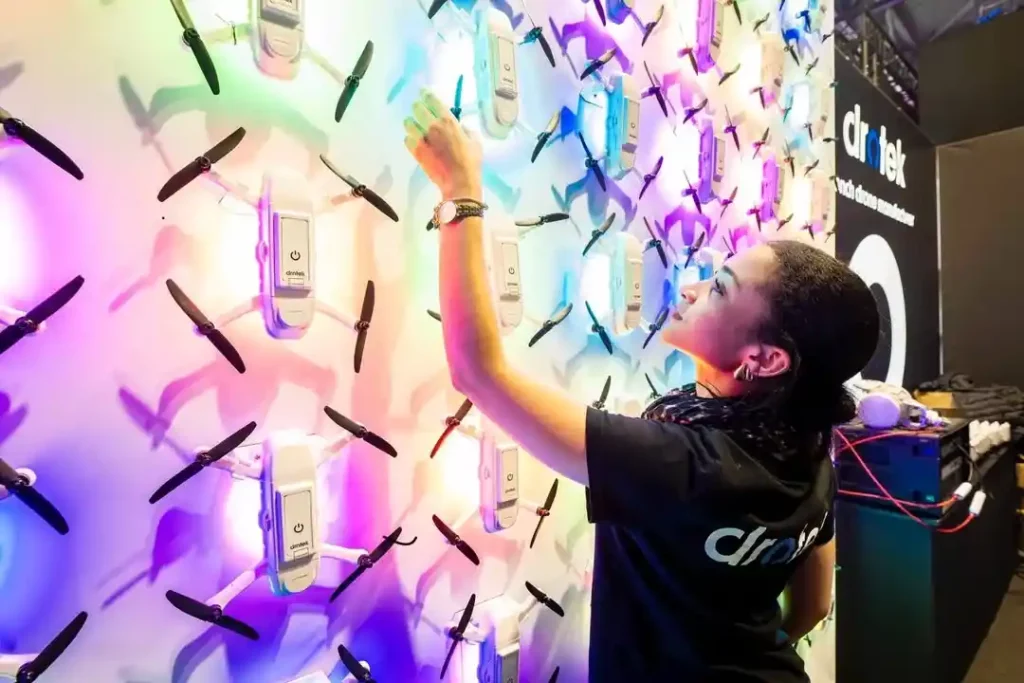 Person interacting with a wall display of multiple small drones mounted in a grid, illuminated by colorful lights at an exhibition booth.