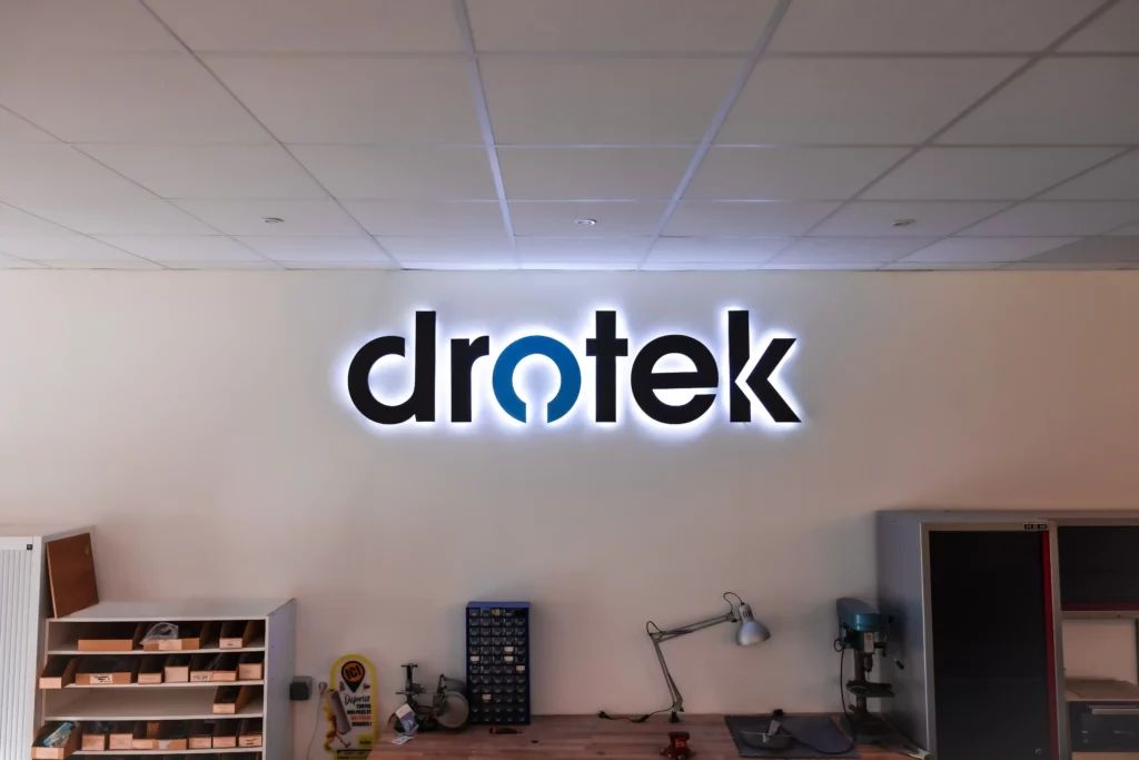 A backlit wall sign displaying the "drotek" logo — bold black lowercase letters with the letter "o" styled as a blue circular symbol with a gap at the bottom, resembling a sensor or connectivity icon — mounted on a white wall in what appears to be an electronics workshop or lab, with shelving, small component organizers, a desk lamp, and a drill press visible in the background.