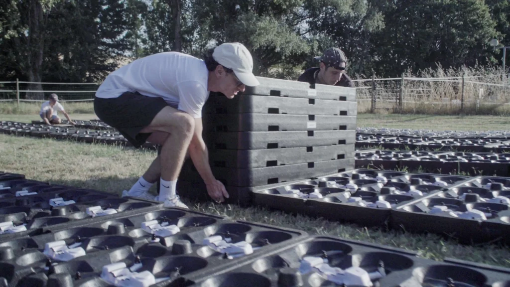 Two Drotek team members lifting stacked drone landing pad trays in a grass field, preparing the launch grid for a drone light show setup.